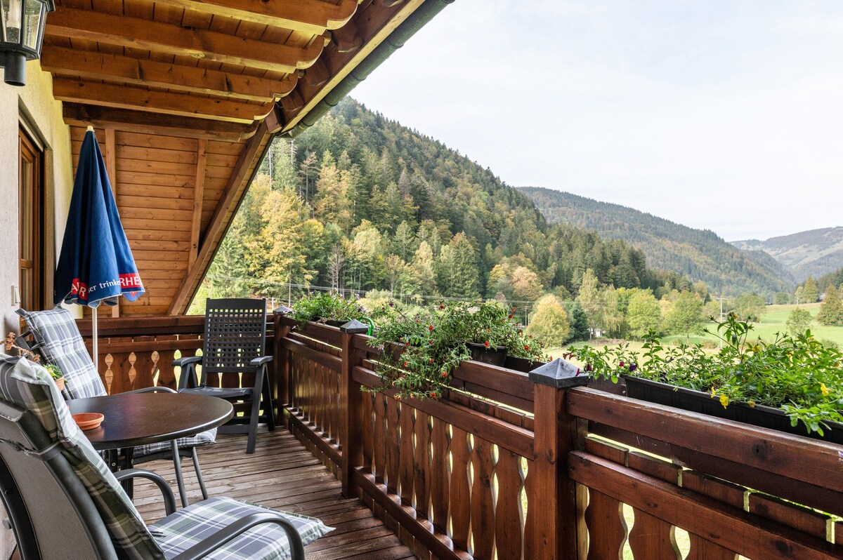 A spacious balcony is featured, showcasing wooden railing adorned with greenery and flowers. A round table and black chairs are positioned for seating. A blue umbrella stands ready for shade, while a backdrop of lush green hills and trees stretches into the distance.