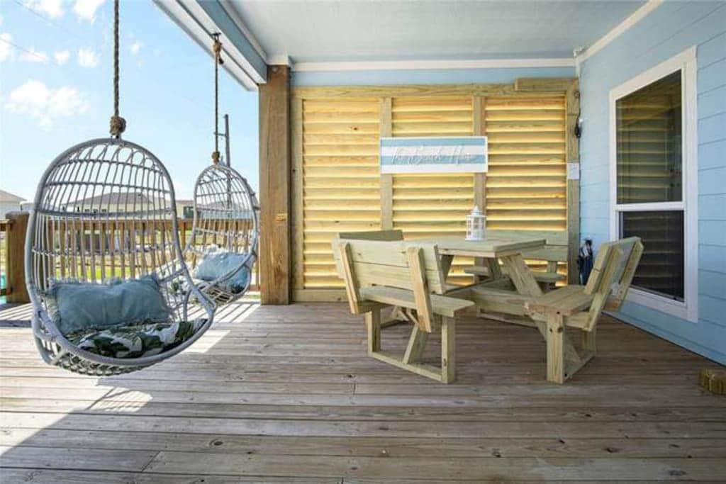 An outdoor area features two hanging chairs and a wooden picnic table, all set on a spacious wooden deck. Sunlight filters through a slatted privacy screen, creating a relaxed atmosphere. The blue exterior wall complements the welcoming design.