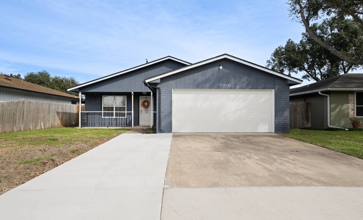 The exterior of a single-story home is depicted, featuring dark blue siding and a welcoming front porch. A well-maintained driveway leads to a garage door, with a neatly landscaped front yard and a fence surrounding the property. Clear skies are visible in the background.