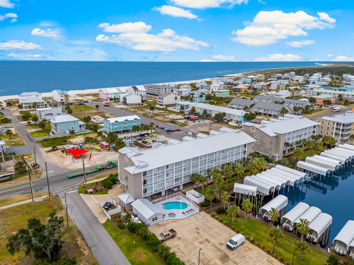 An aerial view captures the coastal landscape of Mexico Beach, featuring a contemporary condo complex near the water. The scene includes a pool area, surrounding palm trees, and a row of covered boat slips. The shoreline is visible in the distance, alongside colorful beach homes.