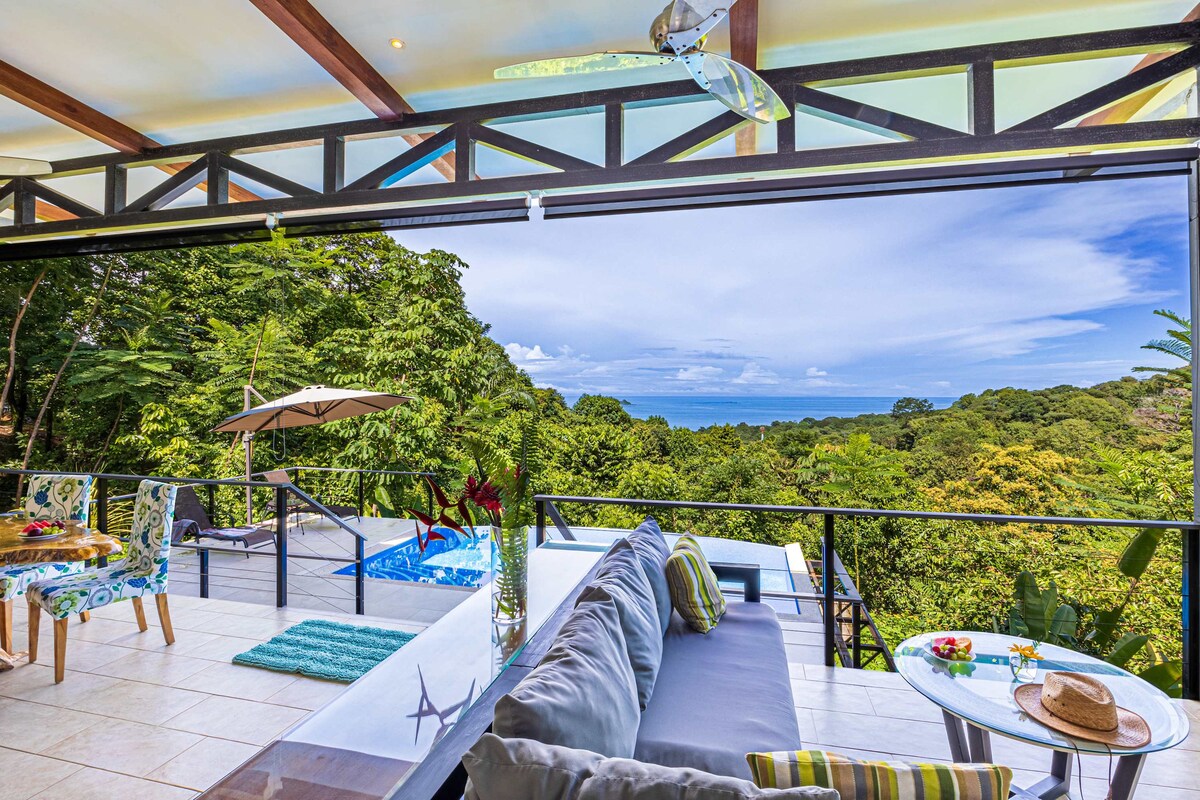 An open living space features a comfortable gray sofa adorned with colorful cushions, offering views of lush greenery and the ocean beyond. A dining area with vibrant chairs is set nearby, while an infinity pool glimmers in the sunlight against the jungle backdrop.