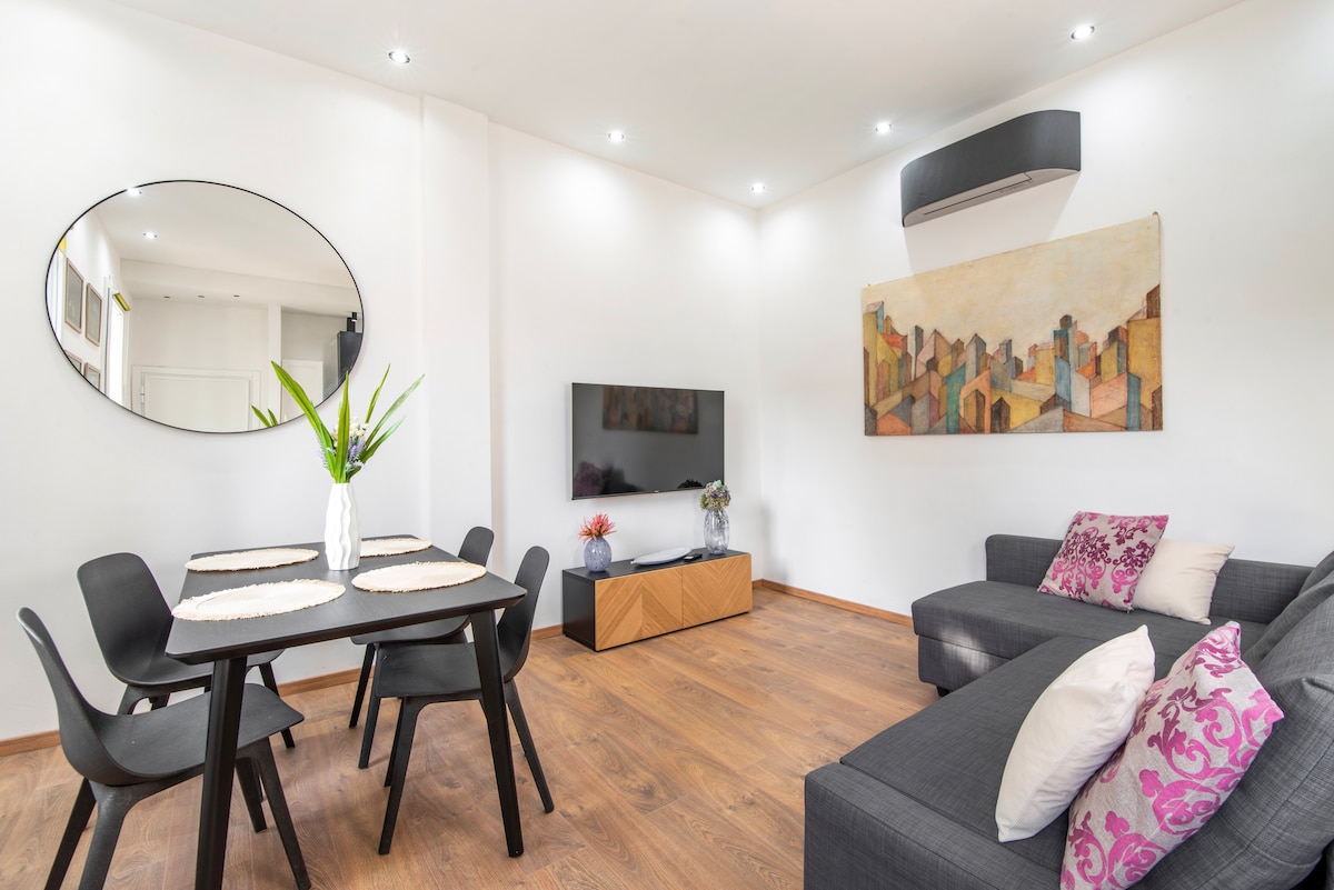 A modern living area features a comfortable dark sofa and a sleek television unit beneath an abstract wall art piece. A dining table is set for four, complemented by black chairs and a decorative plant centerpiece. Soft lighting enhances the open space.