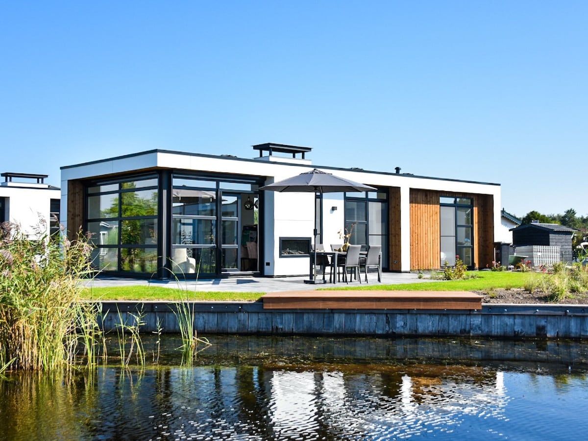 A modern holiday home is situated on the edge of water, featuring large glass windows and a wooden deck. Outdoor furniture includes a table and chairs arranged under an umbrella. The surrounding landscape includes greenery and peaceful water views.