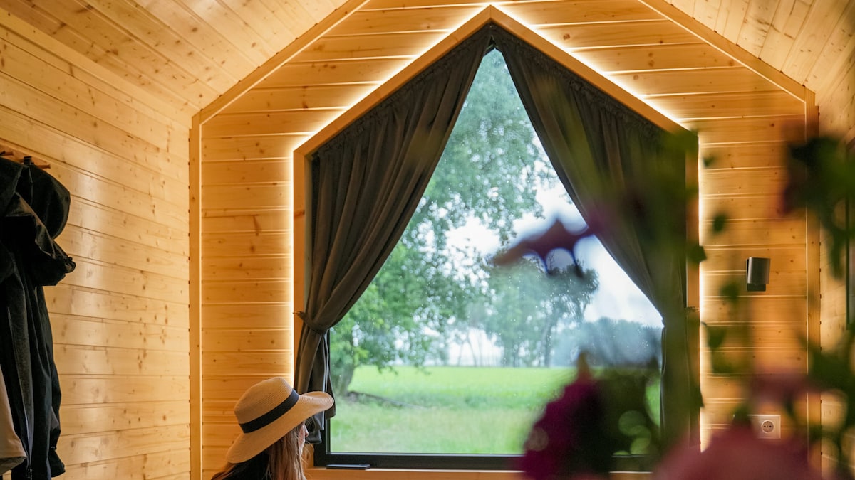 A large, triangular window framed by dark curtains offers a view of the lush green landscape outside. The walls are lined with warm wood panels, creating a cozy and inviting ambiance. A woman in a hat is positioned near the window, adding a sense of scale to the space.