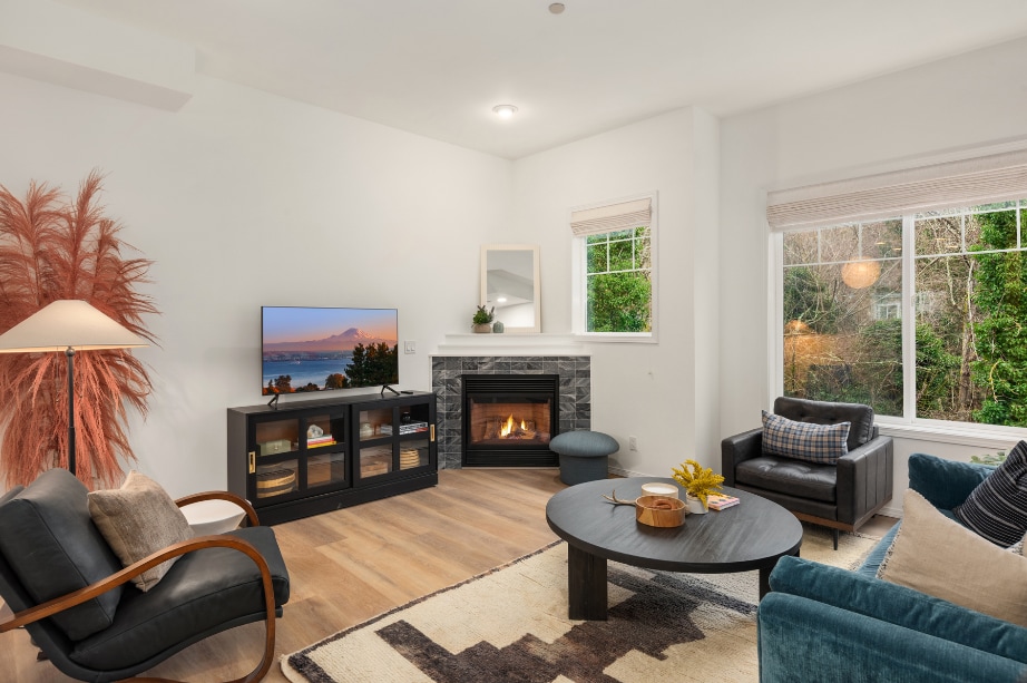 A cozy living area features a fireplace surrounded by decorative stone, complemented by low-profile furniture. A large window allows natural light to fill the room, while a television is positioned on a dark console. Soft textures and a warm color palette create an inviting space.