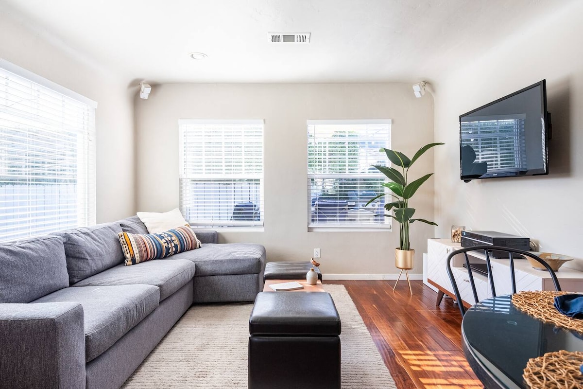 A spacious living room features a gray sectional sofa adorned with decorative pillows. Natural light flows through multiple windows, illuminating the wooden flooring. A mounted TV is positioned on the wall opposite a low white entertainment unit, complemented by a tall indoor plant.