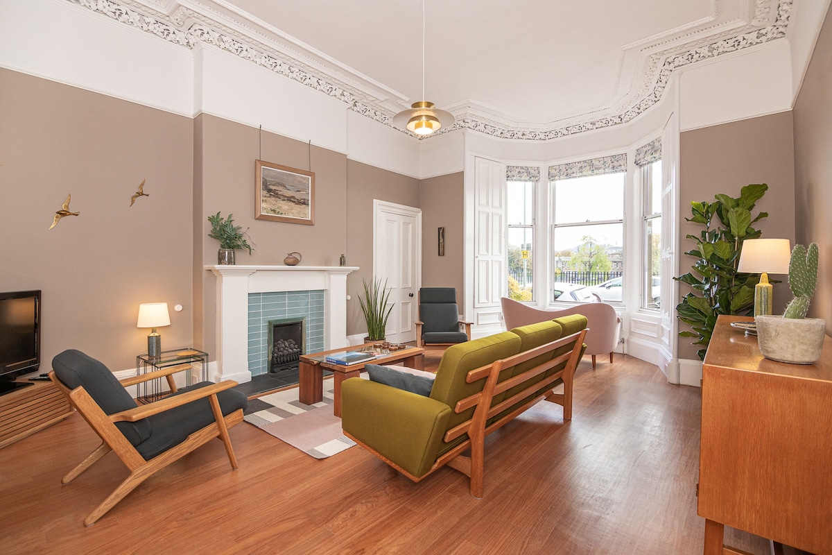 The living area features high ceilings adorned with traditional cornicing. A green sofa and two armchairs are arranged around a coffee table. A decorative fireplace is centered on a light blue tiled wall. Natural light is received through large bay windows, enhancing the space.