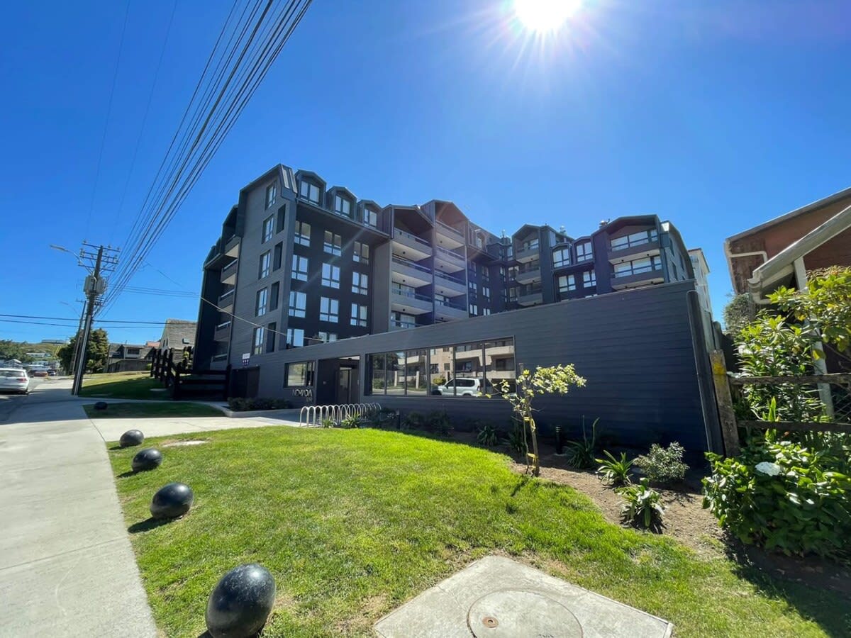 The exterior of a modern building is displayed, showcasing multiple balconies and large windows. A neatly landscaped area features greenery and round black stones along the pathway. Clear blue skies and bright sunlight enhance the welcoming atmosphere.