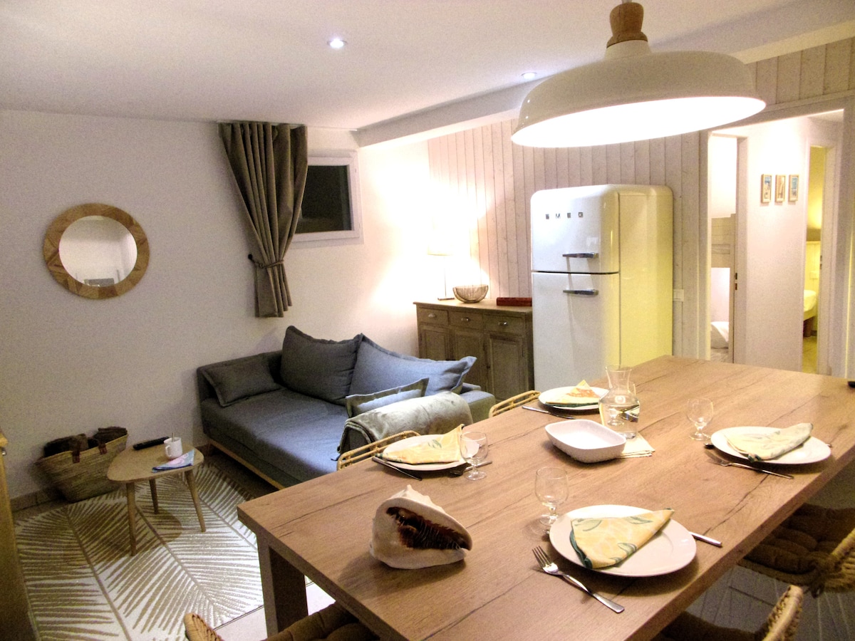 The living area features a comfortable seating arrangement beside a dining table set for four. A beige couch with soft cushions offers a relaxed spot, while an elegant round mirror and refrigerator add charm to the decor. Neutral tones and soft lighting create a cozy ambiance.