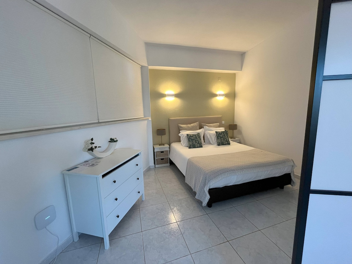 A well-lit bedroom features a spacious bed dressed in neutral linens. Two bedside tables with lamps flanking the bed provide additional lighting. A white dresser is positioned against the wall, and natural light enters through a partially covered window.