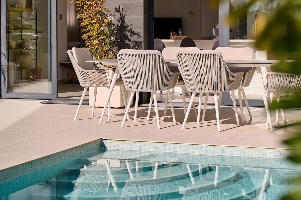 A casual dining area is depicted by a table surrounded by six light-colored chairs. The space is adjacent to a private pool, with reflections of the water visible on the tiled surface. Green plants frame the scene, enhancing the serene ambiance.