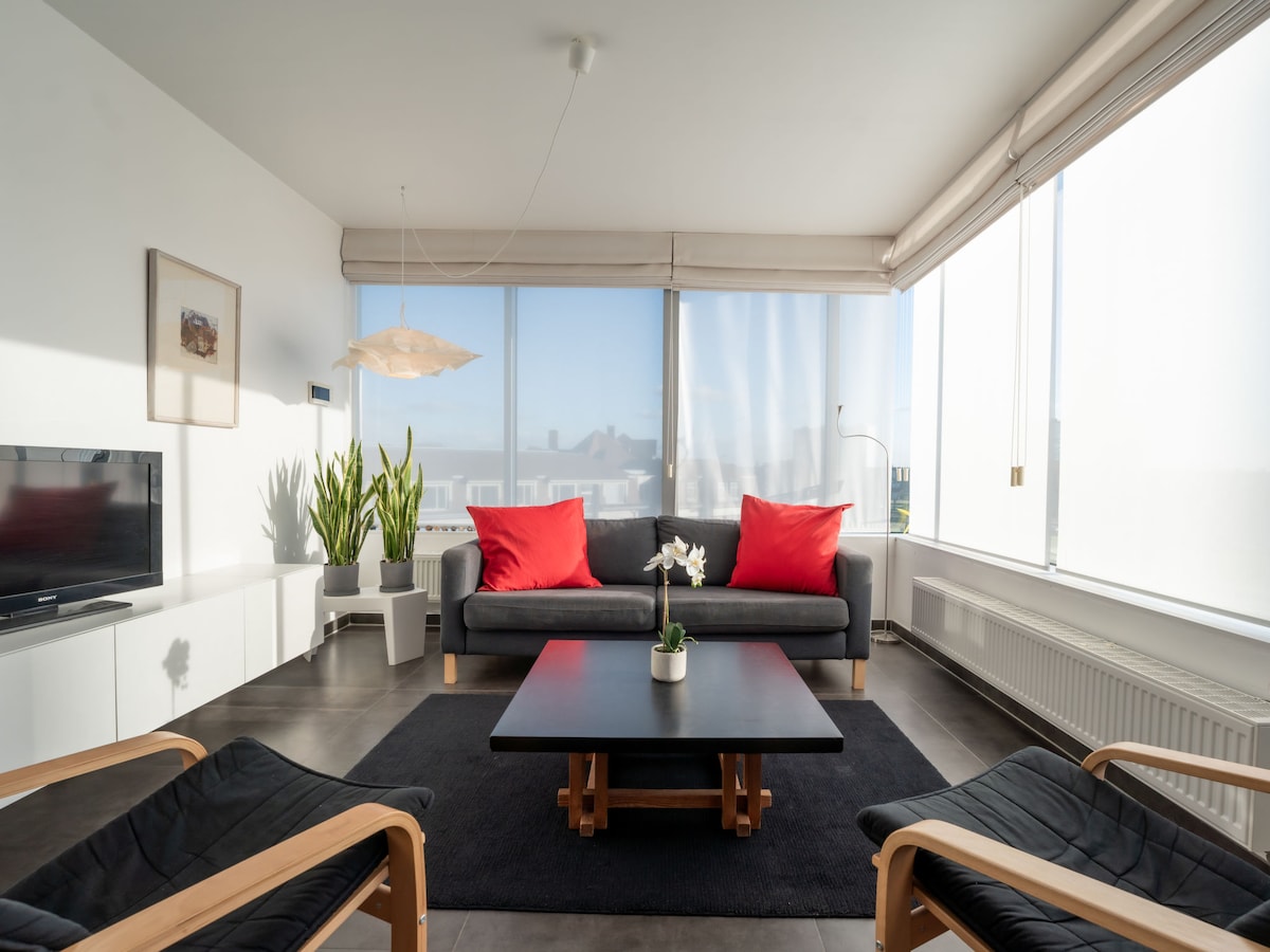 The living area features large windows allowing natural light to fill the space. A modern gray sofa is adorned with two red cushions, complemented by a black coffee table. A light fixture hangs above, and potted plants add a touch of greenery.