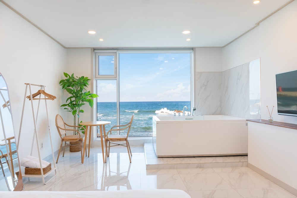A modern open-plan room features a large window offering uninterrupted views of the ocean. A cozy seating area includes a small table and two chairs beside a minimalist wooden surface. A stylish freestanding bathtub adds a touch of elegance to the space.