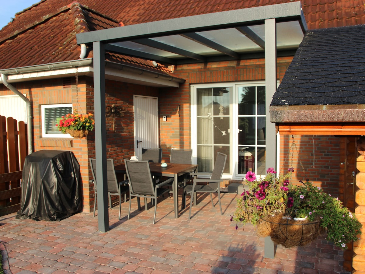 A shaded terrace area is shown, featuring a wooden table surrounded by six chairs. Vibrant flower arrangements hang from the pergola above, and a grill is positioned nearby, creating an inviting outdoor space for gatherings.