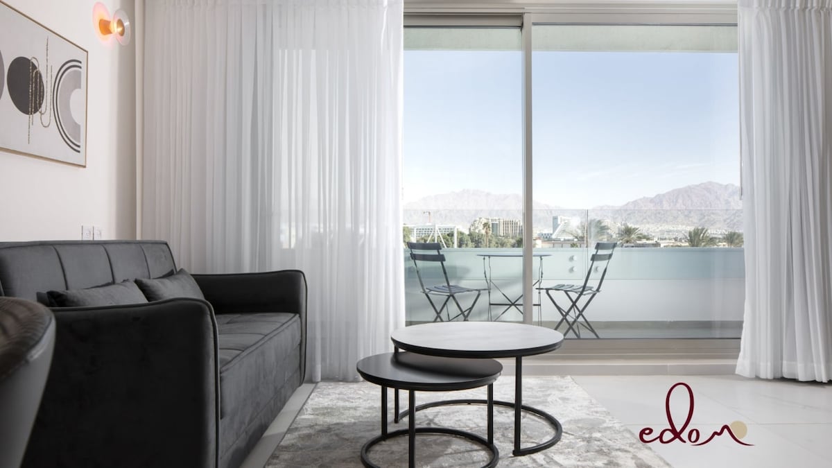 A comfortable seating area is featured with a sleek, dark sofa and a round coffee table. Large glass doors provide a view of the outdoor balcony, which includes two chairs and a small table, framed by light, sheer curtains that filter natural light.