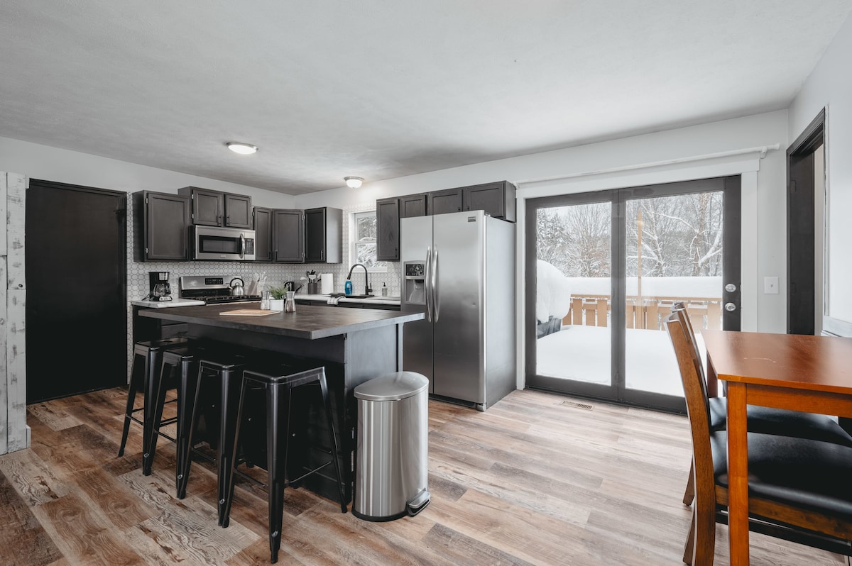 An open kitchen area features dark cabinetry and modern stainless steel appliances. A central island with seating accommodates four. Sliding glass doors provide access to a winter scene outdoors, while a dining table for four offers additional space for meals.