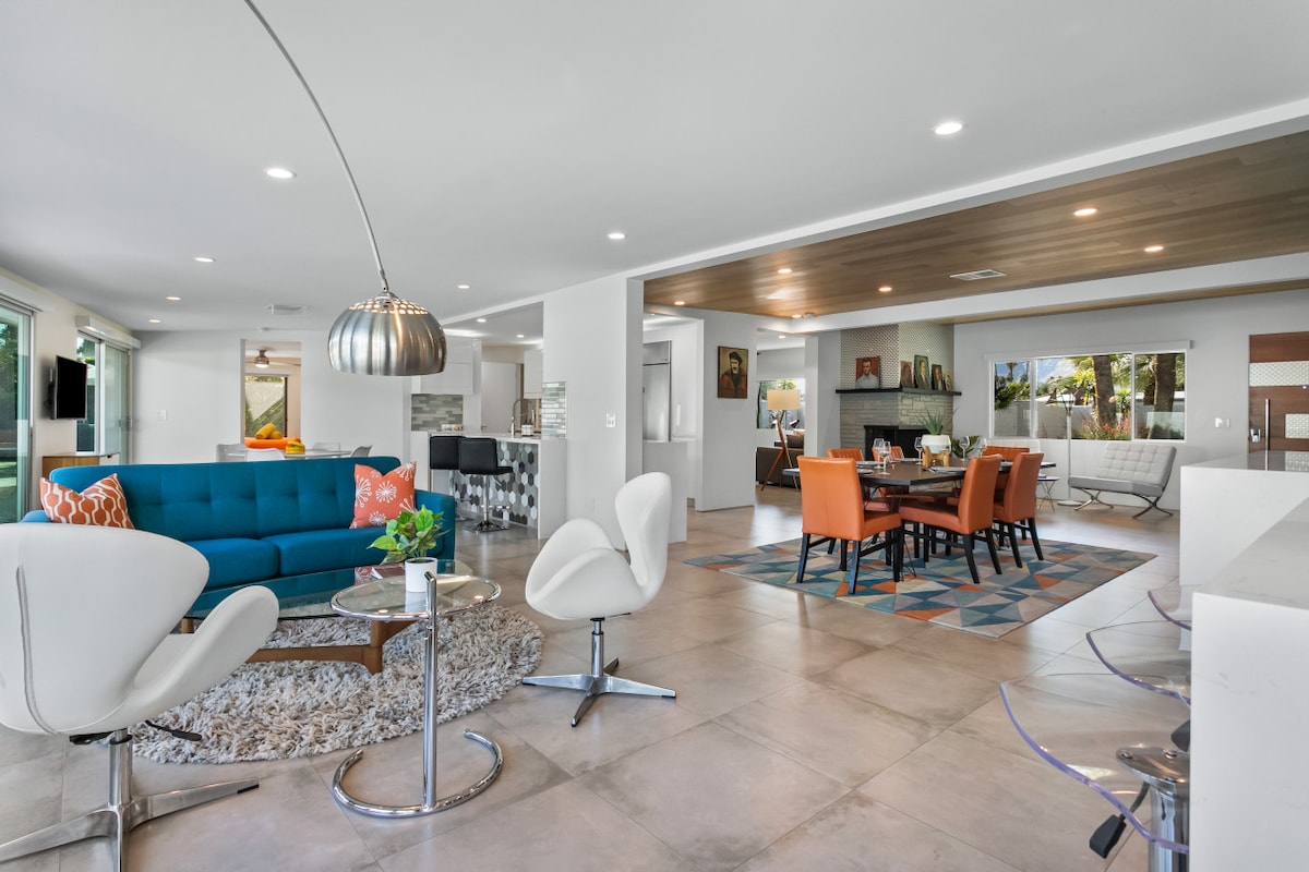 An open living area showcases a vibrant blue sofa paired with white accent chairs and a unique coffee table. Natural light flows in through large glass doors, illuminating the stylish dining space featured with warm orange chairs and a patterned rug.