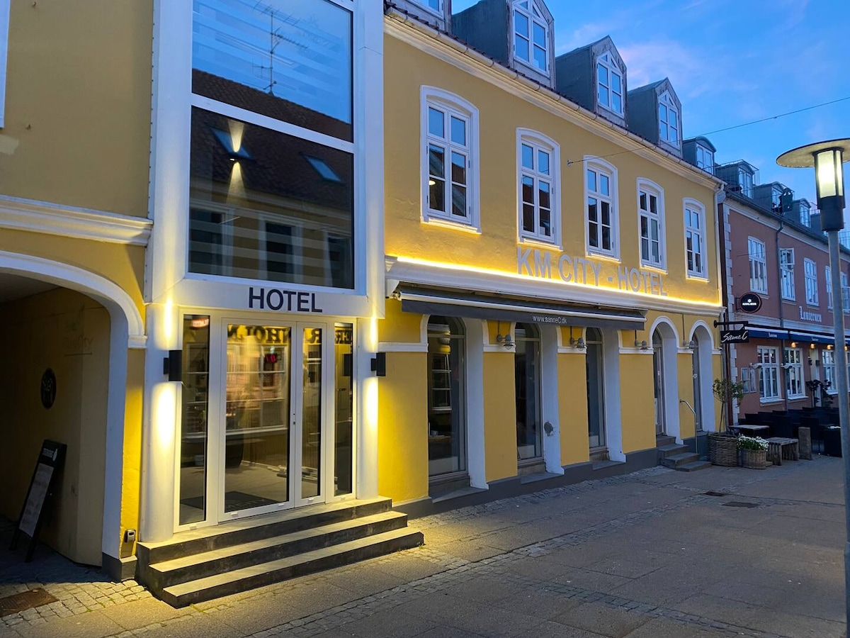 A modern hotel facade is illuminated in soft light, highlighting a welcoming entrance with steps. The building features yellow exterior walls and large windows, offering an inviting atmosphere. Nearby storefronts and outdoor seating can be seen along the pedestrian street.
