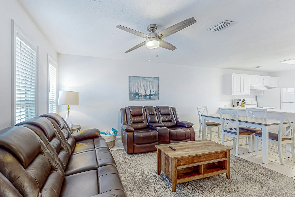 A spacious living area features two comfortable leather sofas and a wooden coffee table. Natural light enters through window blinds, highlighting a nautical-themed painting on the wall. The open layout connects to a dining area with a light-colored table and chairs.