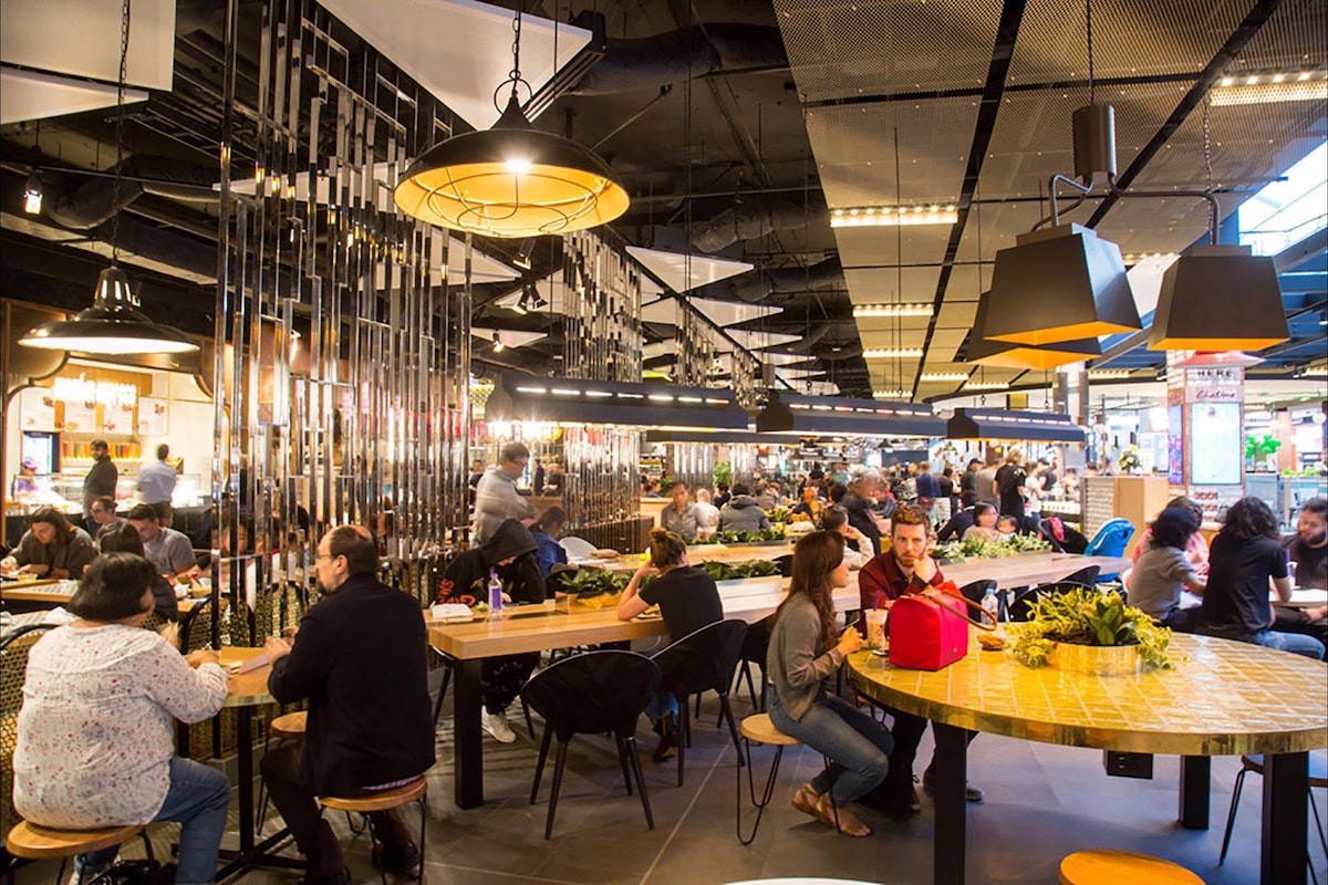 A lively dining space is filled with patrons enjoying various meals. Large tables with light-colored tops and stylish chairs are scattered throughout. Pendant lights hang from the ceiling, casting warm illumination, while the atmosphere buzzes with conversation in this modern, open layout.