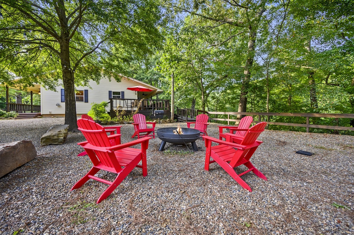 Four red Adirondack chairs surround a central fire pit, set on a gravel area shaded by trees. A cabin with a red-roofed porch is visible in the background, enhancing the natural surroundings.