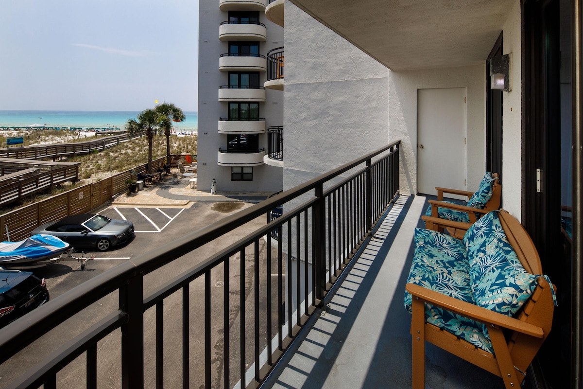 A private balcony is showcased, featuring two wooden chairs with vibrant tropical cushions. The view extends to the beach and the Gulf waters, while the parking lot and surrounding area are visible below. The balcony is accessed directly from the interior of the unit.