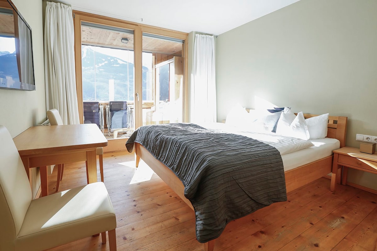 A spacious bedroom features a large bed adorned with crisp white linens and decorative pillows. Natural light fills the room through large glass doors leading to a balcony. A simple wooden table and light-colored chair complement the warm wooden floor.