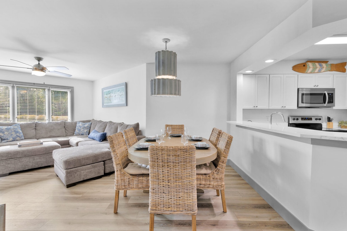 A bright and airy dining area features a round table surrounded by woven chairs. A modern light fixture hangs above, while a spacious sectional sofa is positioned nearby. A fully equipped kitchen is visible in the background, showcasing stainless steel appliances and a decorative fish accent.