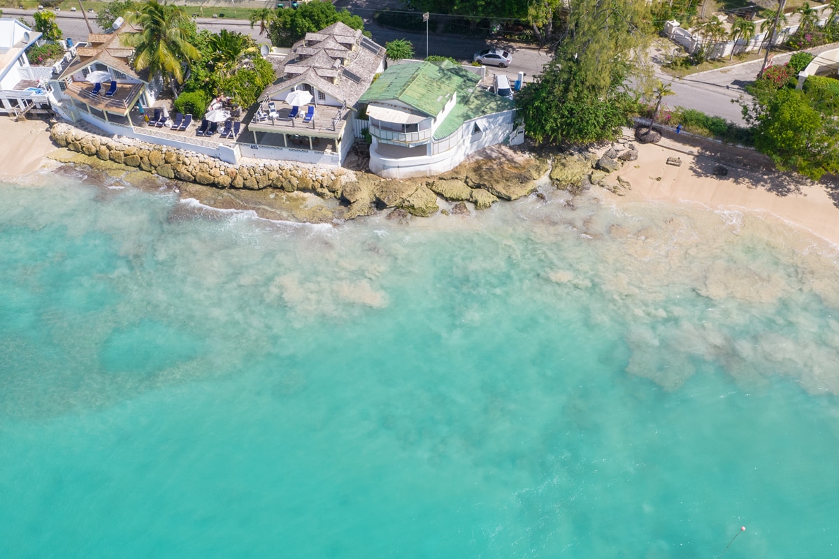 An aerial view showcases the beach villa's proximity to the turquoise waters of Mullins Beach. The shoreline features gentle waves lapping against the rocky edge, with lush greenery and beachside setups visible, enhancing the coastal charm of the property.