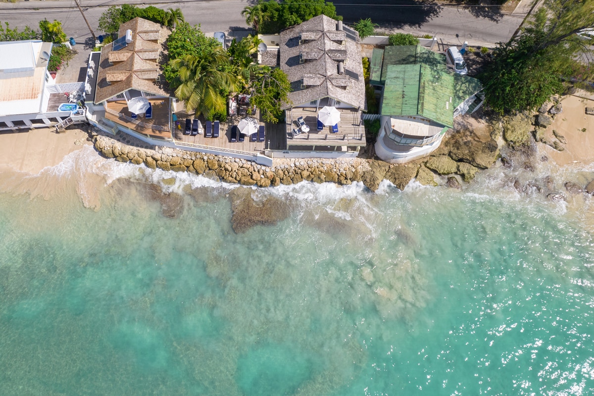 An overhead view showcases the villa's beachfront location with gentle waves lapping at the shoreline. The property features a serene arrangement of palm trees, sun loungers, and shaded sitting areas, set against the backdrop of clear turquoise waters and rocky shore.