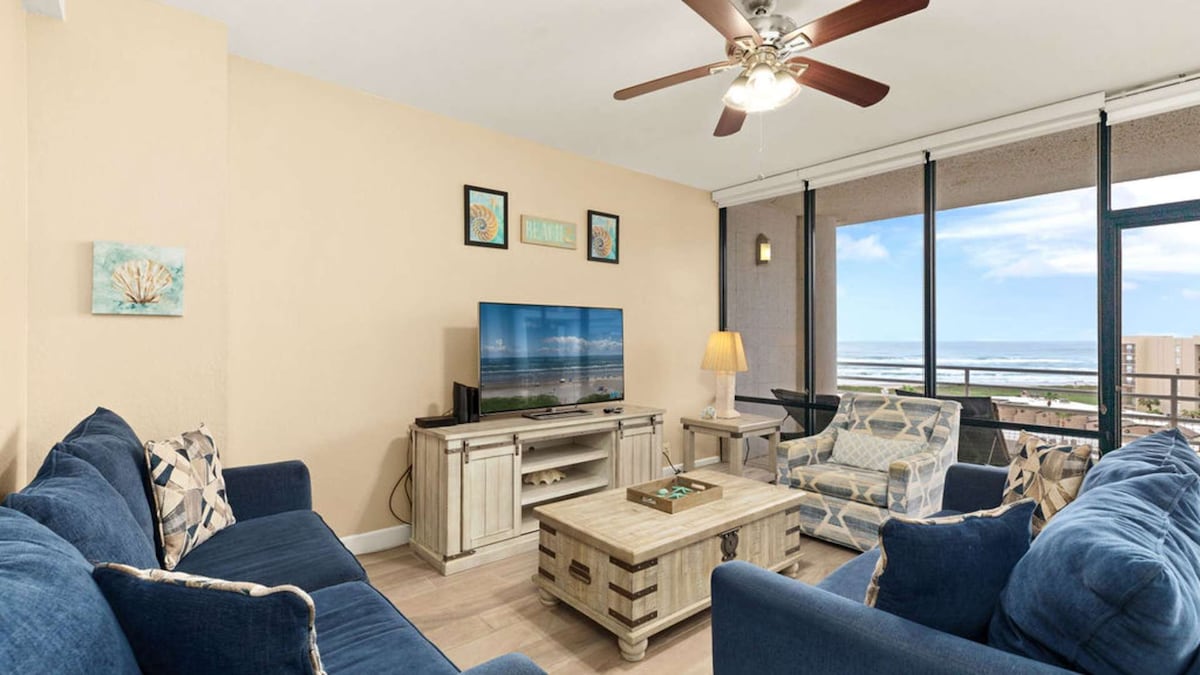 A spacious living area features comfortable seating with blue couches and patterned chairs. A wooden coffee table is centered in front of a large flat-screen TV, which is placed on a light-colored media console. Floor-to-ceiling windows provide expansive views of the coastline.