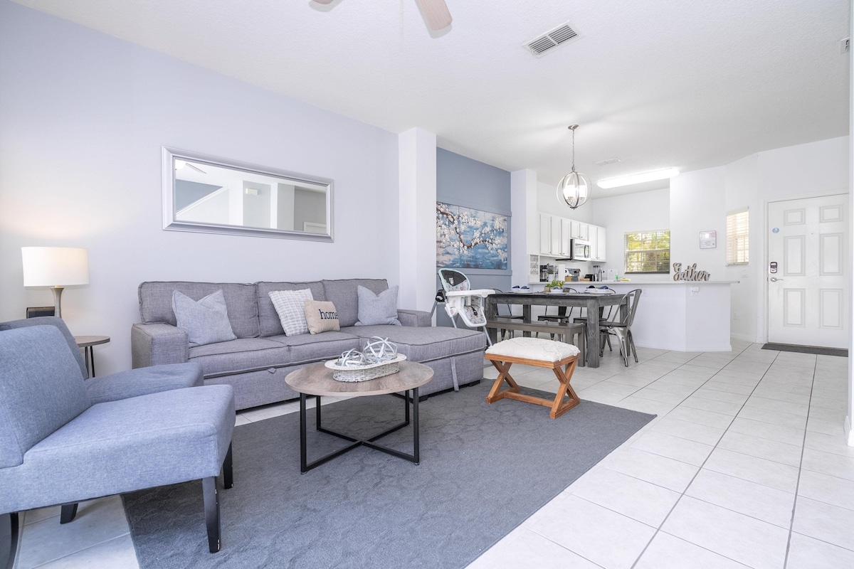 A modern living area features a gray sectional sofa paired with a small wooden table. Natural light fills the space, highlighting a neutral-colored rug and a circular coffee table at the center. The open layout leads to a dining area, visible in the background.