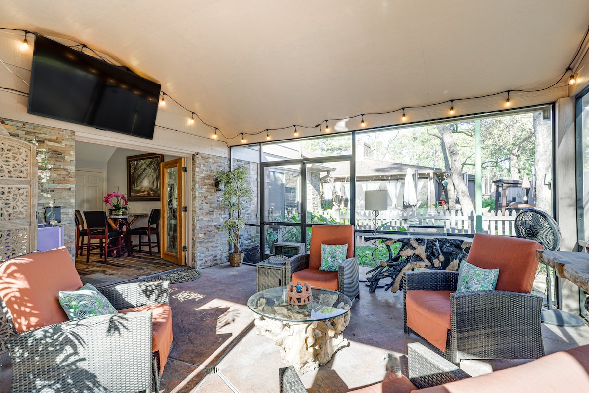 A spacious outdoor living area is enclosed by screens, featuring comfortable seating arrangements with colorful cushions. A coffee table made of stone is centrally placed, and string lights provide subtle illumination. A large wall-mounted TV is positioned above the seating area.