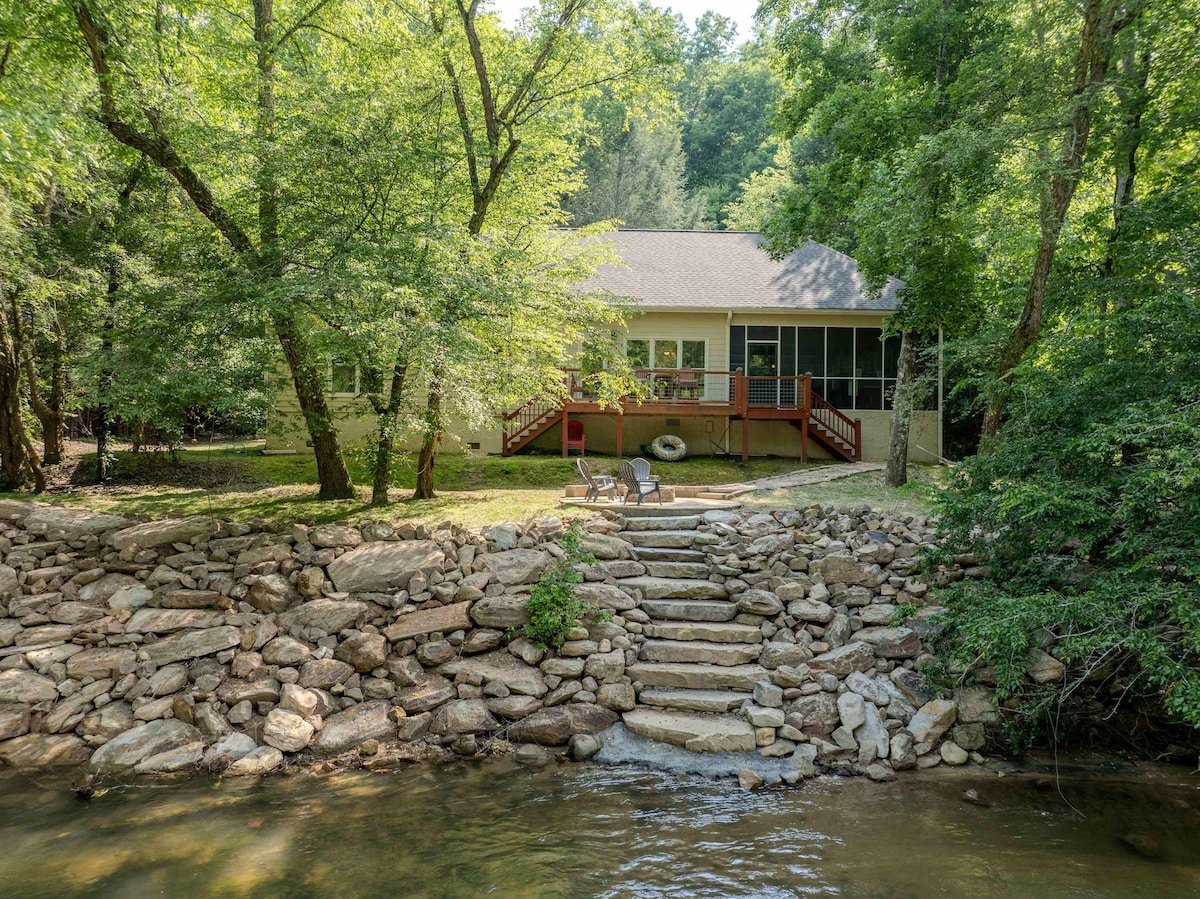 A welcoming vacation rental is set beside the Cartecay River, featuring a stone pathway leading to the water. Surrounded by trees, a screened porch and outdoor seating are visible on the deck, providing inviting outdoor space for relaxation.