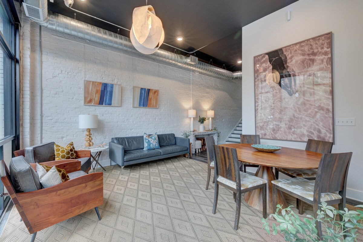 An open living area features large windows, allowing ample natural light. A sleek leather sofa and mid-century modern chairs are arranged around a decorative table. The dining area includes a wooden table with dining chairs, complemented by original wall art and a geometric-patterned rug.