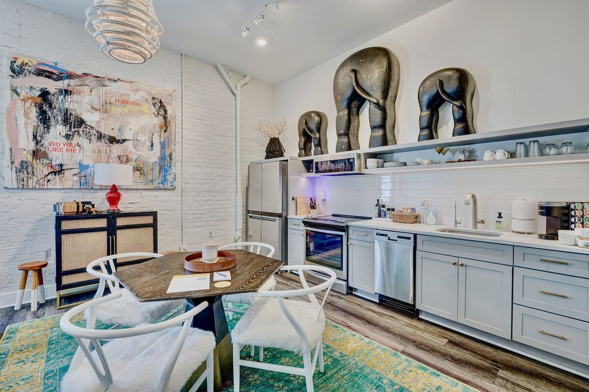 A modern kitchen is presented with stainless steel appliances and sleek cabinetry. A round dining table is surrounded by four white chairs, with a decorative area rug beneath. Artwork featuring elephants adorns the wall, adding a unique touch to the space.