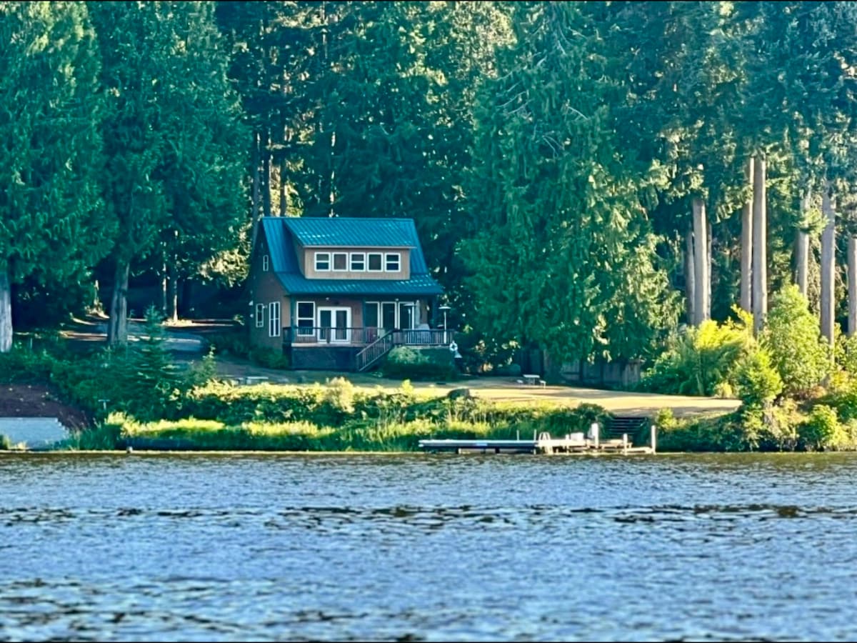 A charming lakefront home is nestled among tall trees, featuring a green metal roof and spacious deck. The property overlooks a serene lake, with a private dock visible in the foreground, creating an inviting scene for relaxation and recreation.