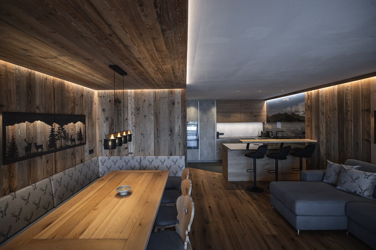 A combined living and dining area features wooden accents and soft lighting. A large wooden dining table is surrounded by upholstered seating. An inviting gray sofa occupies one corner, with a modern kitchen visible in the background, equipped with sleek appliances and bar seating.