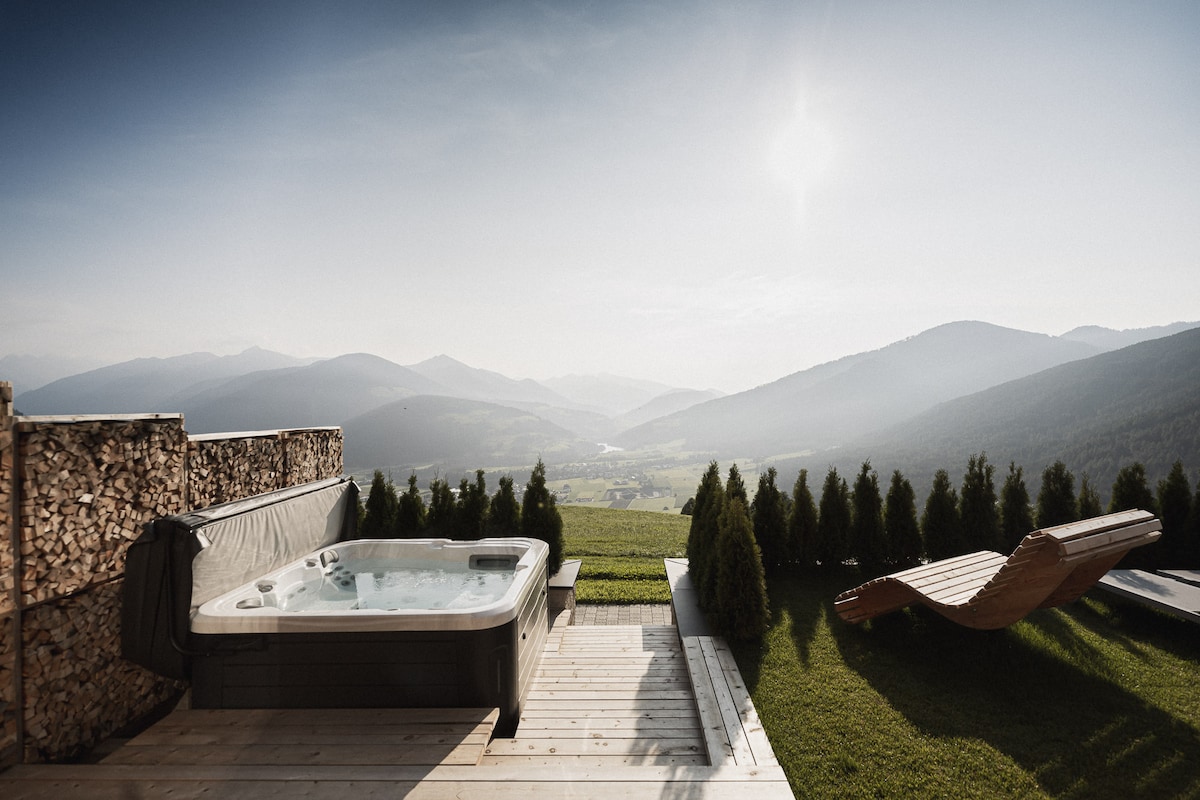 A private outdoor area features a hot tub surrounded by low hedges, with a view extending over a lush valley and distant mountains. A wooden lounge chair rests on the deck, complemented by the serene natural landscape and a clear sky.