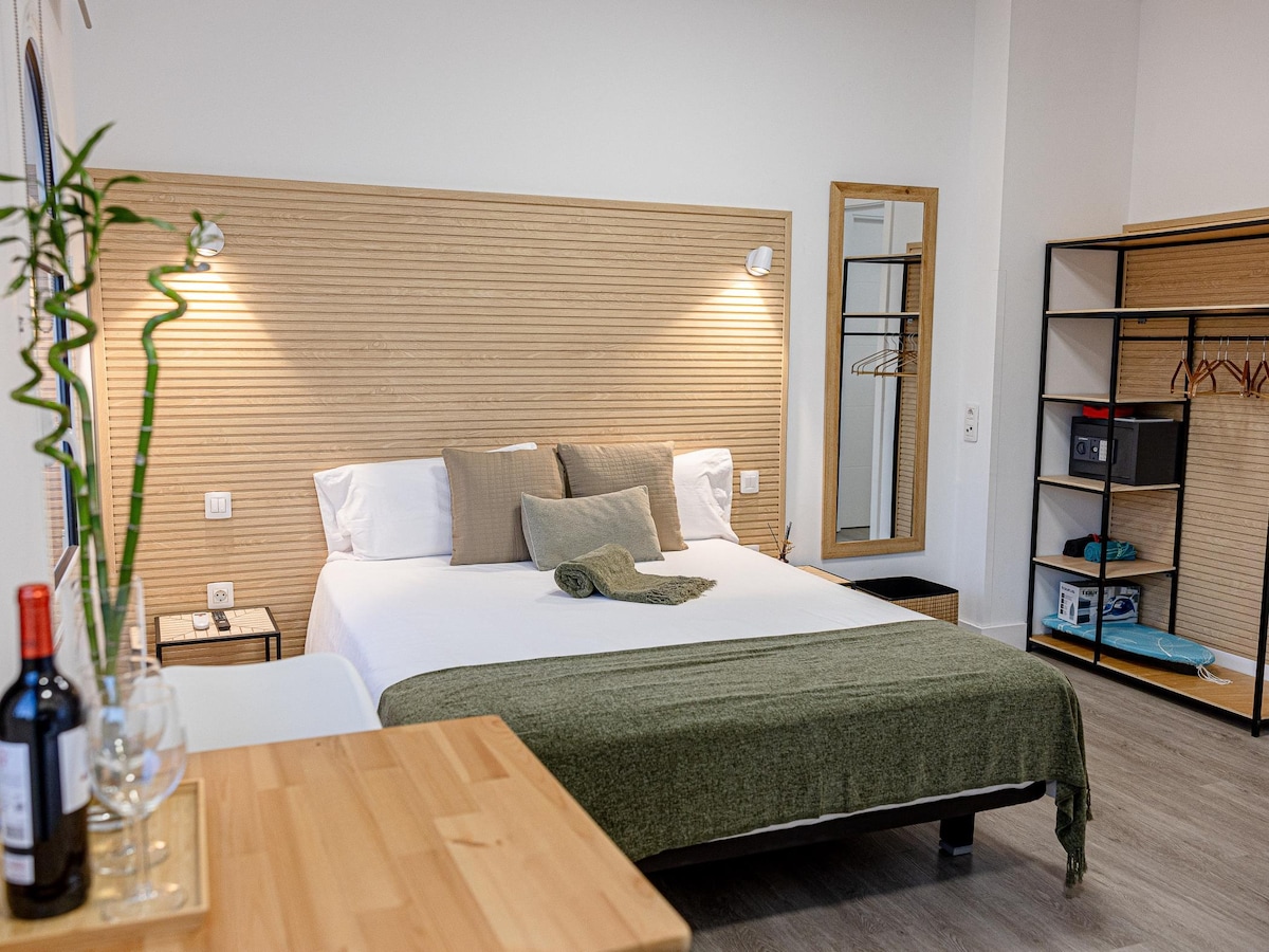 A modern room features a spacious bed with neutral-toned bedding and decorative cushions. A simple wooden headboard complements the light-colored walls. A dining table with wine glasses is positioned nearby. A wardrobe and shelving unit are visible, providing convenient storage options.