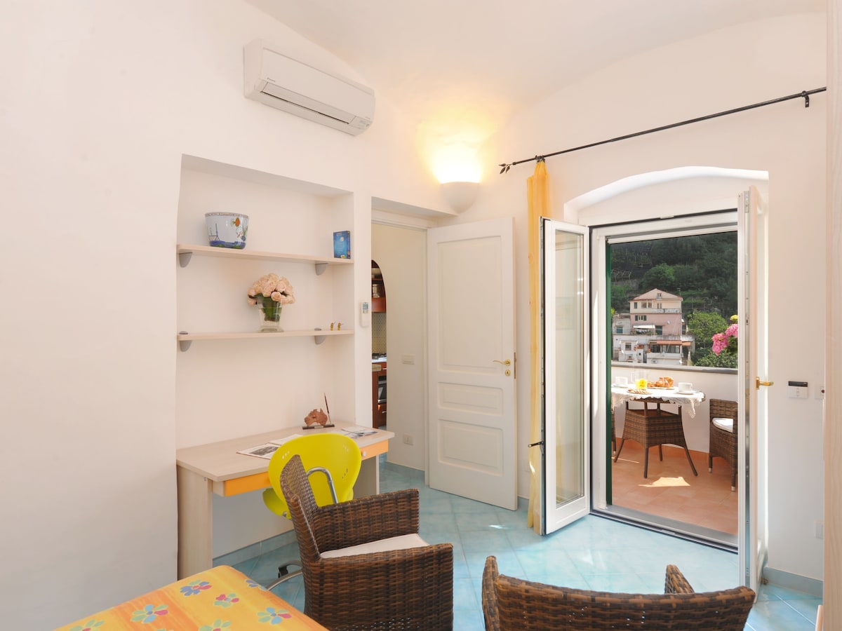 A bright living space features a small desk with a yellow chair and two wicker chairs near a floral bedspread. Natural light enters through the open balcony door, which leads to a terrace with views of the surrounding landscape.