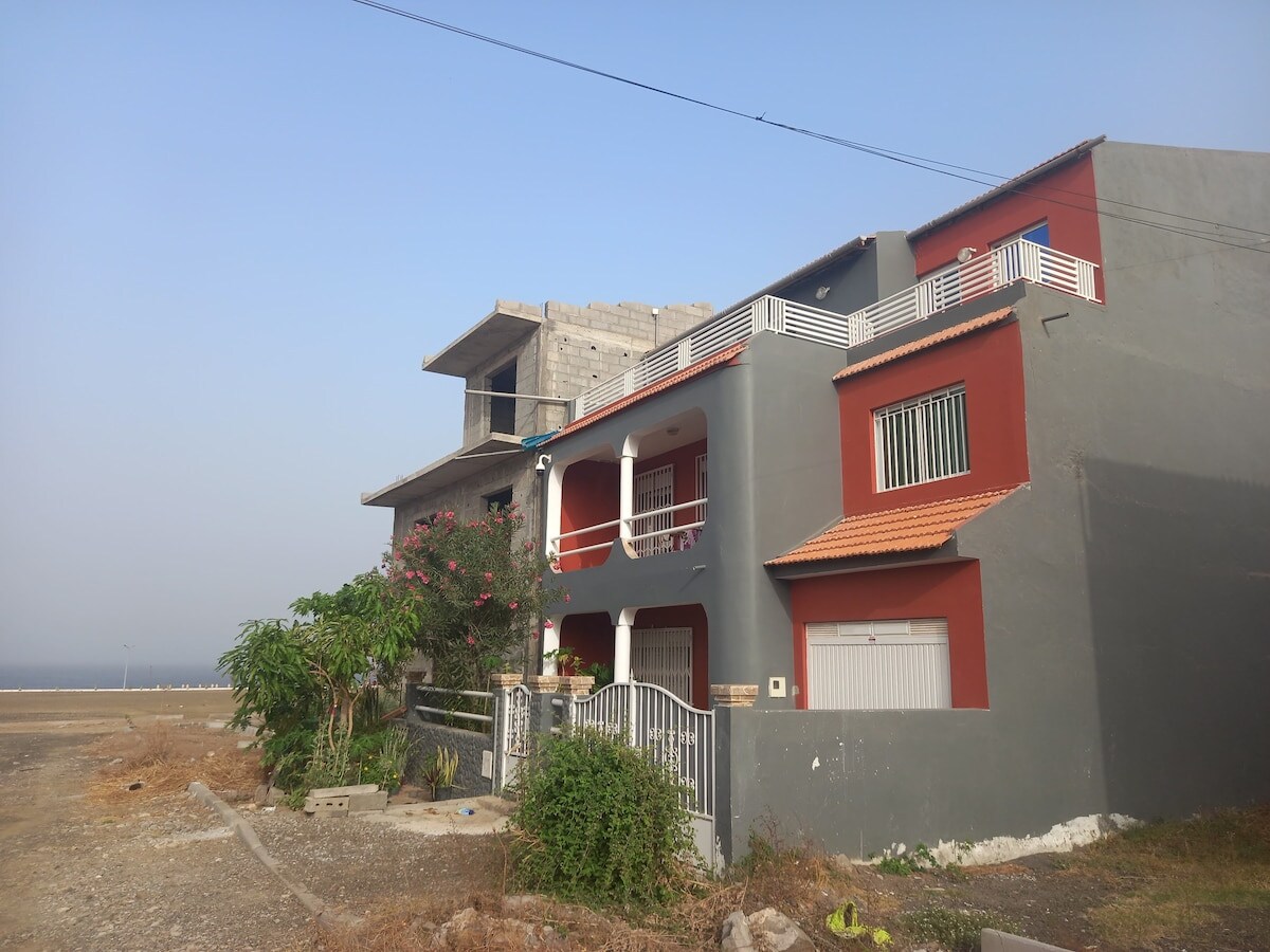 A multi-level building is prominently displayed, featuring a combination of light gray and red accents. Balconies extend from the upper floors, offering views of the surrounding area. Lush vegetation is present near the entrance, enhancing the structure's appeal in a tranquil setting.