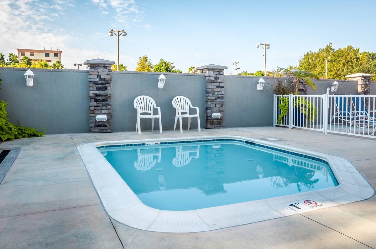 A small outdoor pool is surrounded by a smooth concrete deck, featuring white plastic chairs positioned nearby. The pool reflects a clear blue sky above, while the area is enclosed by a low stone wall and greenery in the background, creating a tranquil setting.