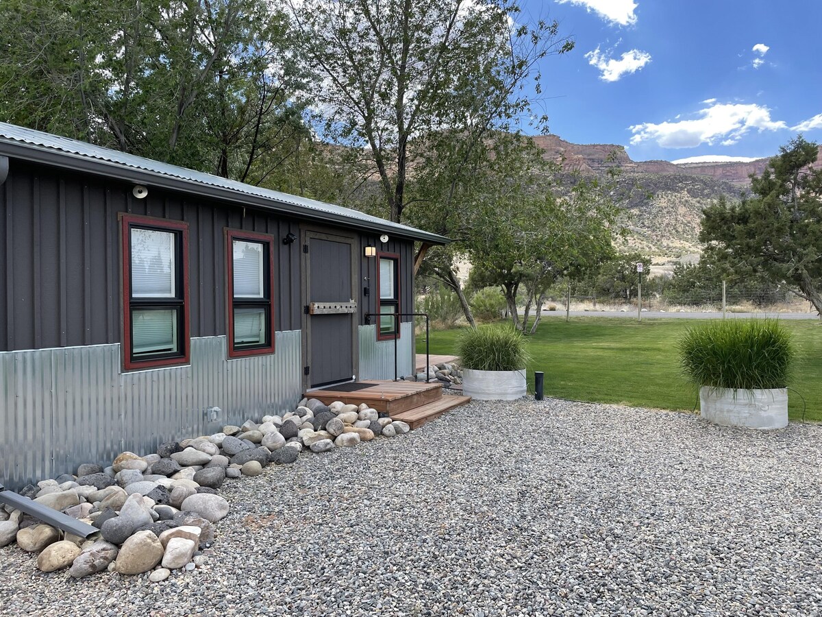 A modern tiny home is set against a natural backdrop, featuring grey siding and large windows in a rich red trim. A wooden porch with steps is approached by a pathway of smooth stones, while green grass and trees surround the property, enhancing the outdoor serenity.