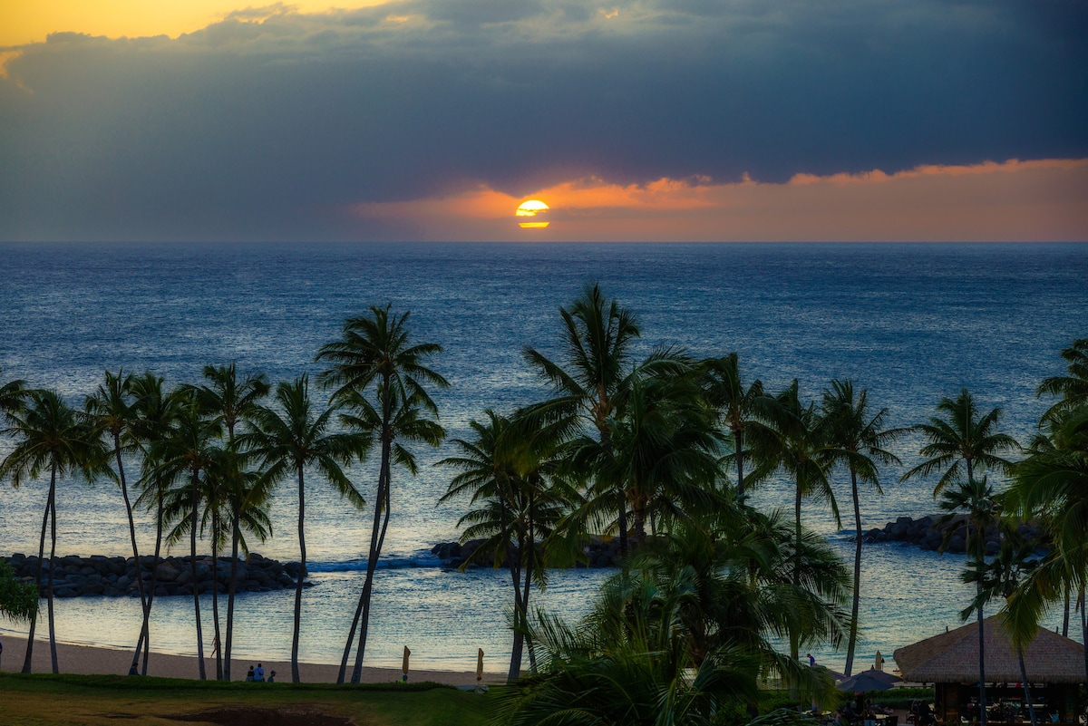 A serene sunset is captured over the ocean, with warm hues of orange and pink blending into the horizon. Silhouetted palm trees frame the scene, while gentle waves lap against the shoreline, creating a peaceful atmosphere at the resort.
