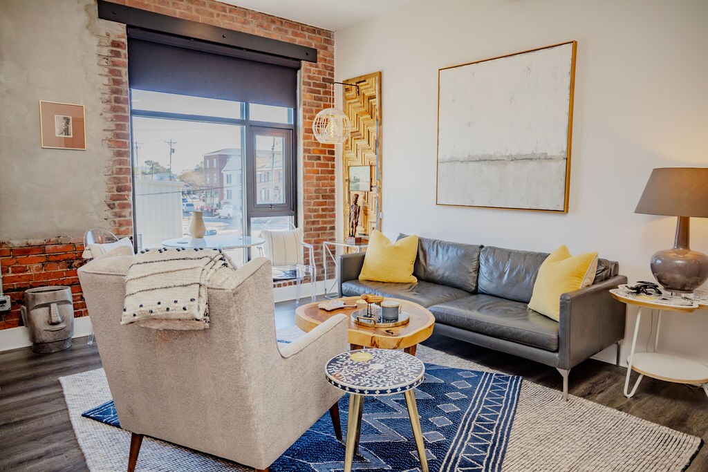 A cozy living area features an inviting leather sofa adorned with yellow accent pillows. An elegant coffee table is centered on a patterned area rug, while a large window allows natural light to fill the space. Exposed brick walls complement modern artwork and decor elements.
