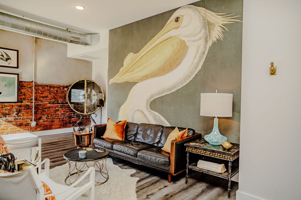 A stylish living area is showcased with a leather sofa facing a circular mirror. A vibrant mural of a pelican adorns the wall, complementing the exposed brick. A low coffee table is placed on a soft area rug, accented with decorative pillows and a lamp.