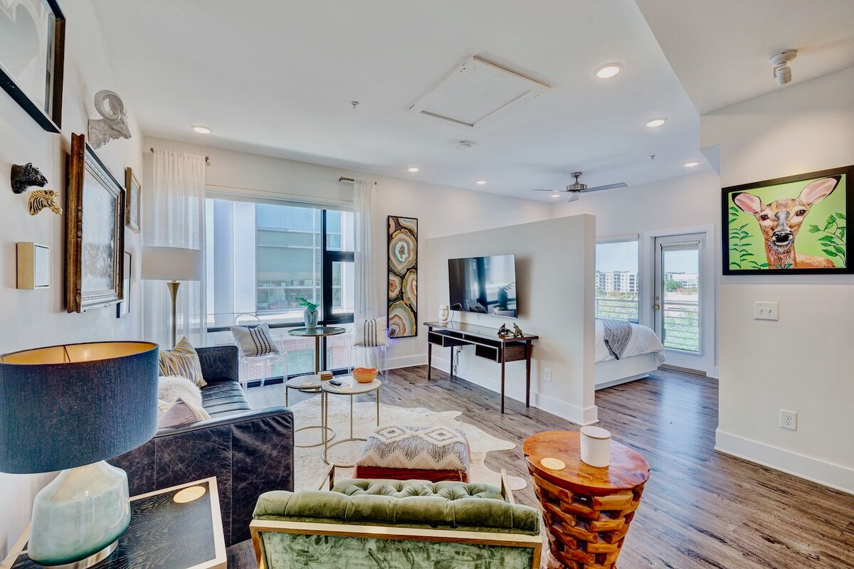 A modern living area showcases a comfortable seating arrangement with a large television mounted on the wall. Natural light filters through expansive windows, highlighting decorative art pieces. A round dining table complements the space, while a glimpse of a cozy sleeping area is visible through the door.