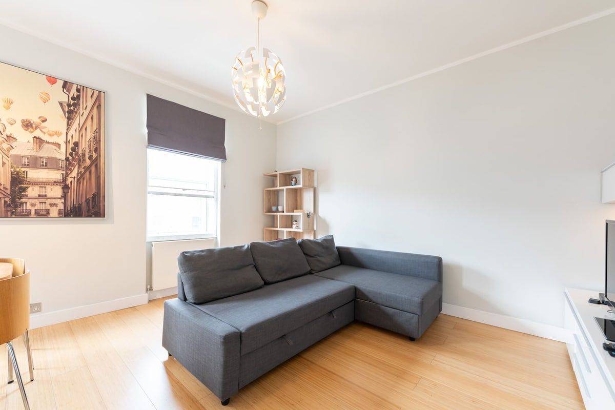 A modern living area features a spacious sectional sofa in a neutral tone. A stylish light fixture hangs from the ceiling, while a piece of wall art depicting a street scene adds character. Sunlight streams through a window covered by a light roller shade.