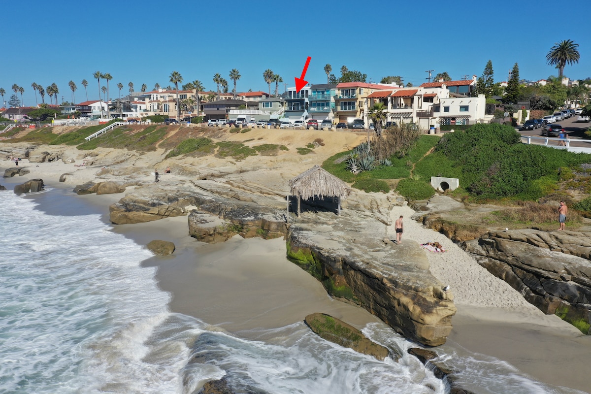 A panoramic view captures the oceanfront property situated above the beach, with gentle waves lapping at the sandy shore. Nearby, a rocky outcrop is visible, along with residential buildings and palm trees lining the coastline under a clear blue sky.